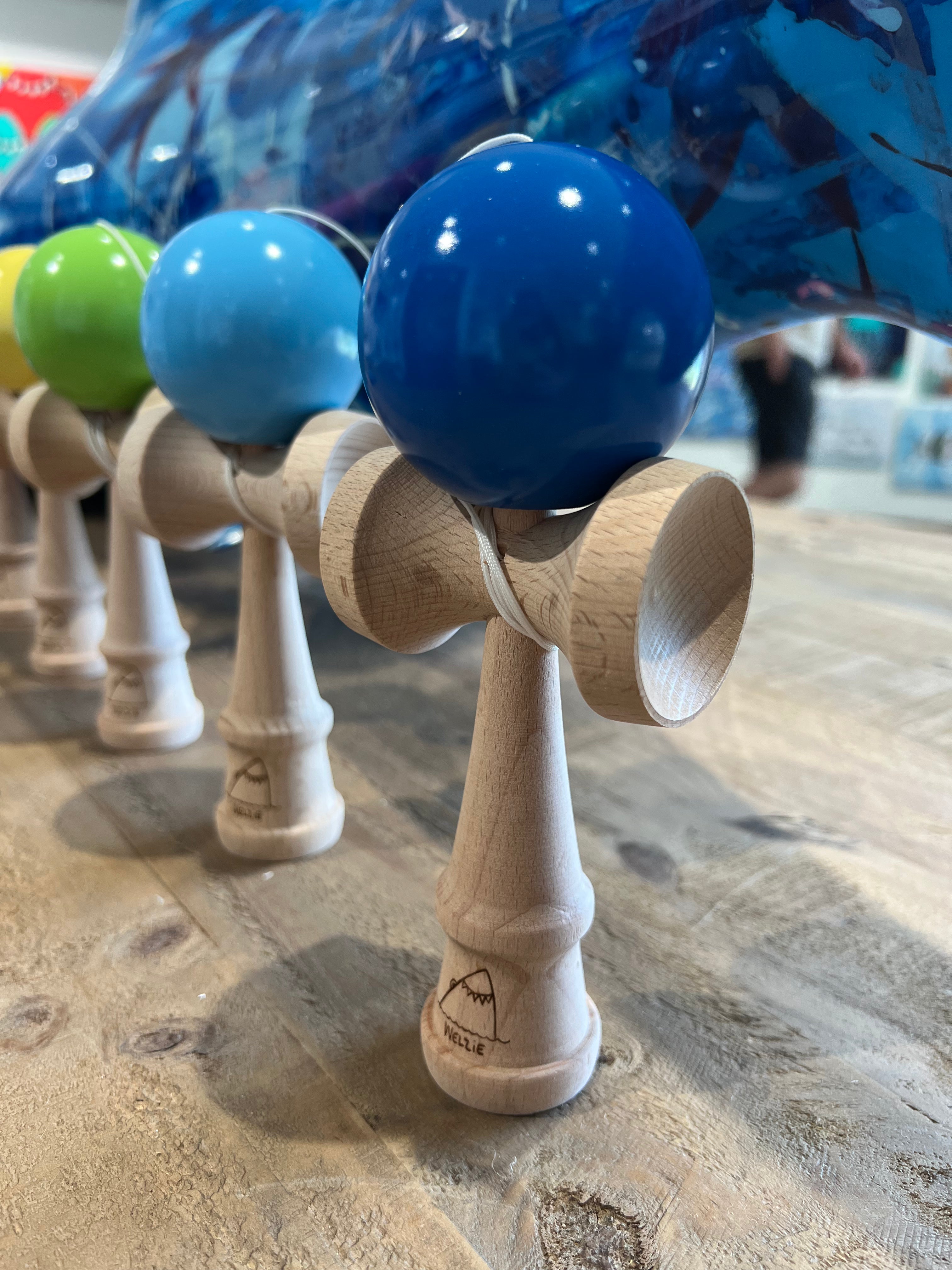 A close-up of several wooden kendama with colorful spherical heads, including blue, green, and light blue, arranged on a wooden surface.