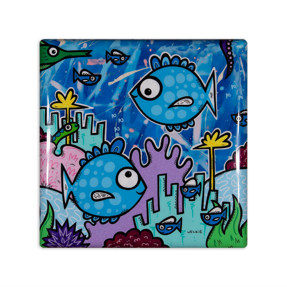 A square glass art tile featuring a colorful cartoon underwater scene with blue spotted fish, coral, and sea plants on a white background.