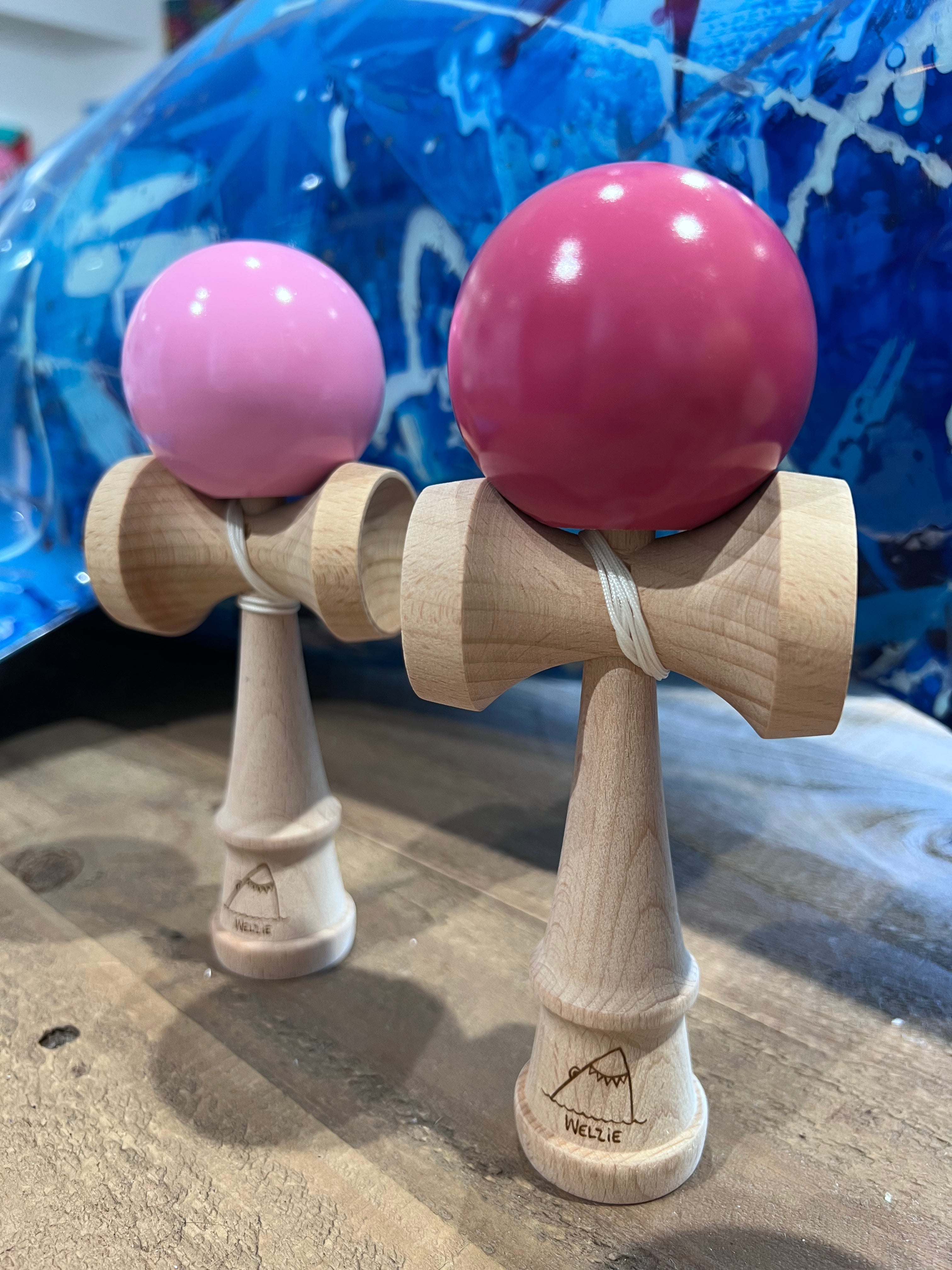 A close-up of several wooden kendama  with colorful spherical heads, including Pink and Dark Pink arranged on a wooden surface.