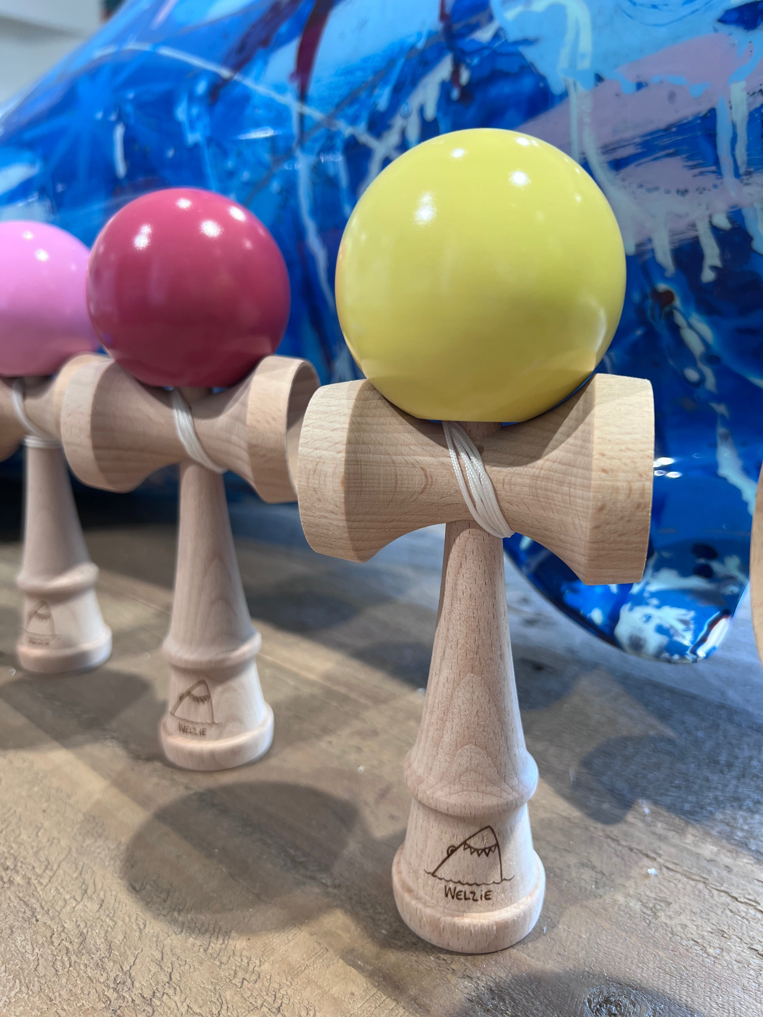 A close-up of several wooden kendama balls lined up in a row, with the frontmost ball in bright yellow and the next in pink.